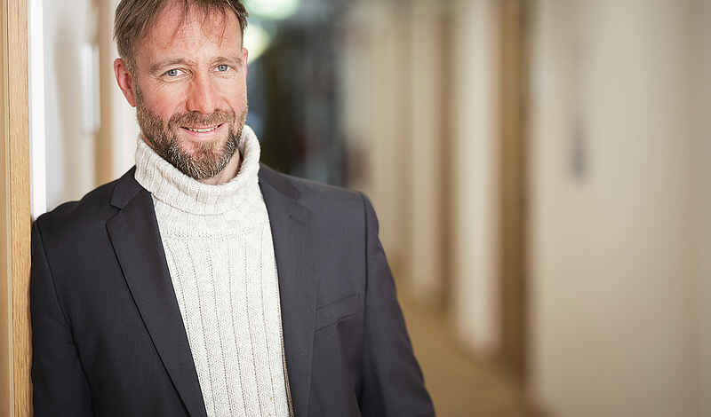 Ein Mann mit kurzen braunen Haaren und Bart lehnt lächelnd seitlich an einer Wand. Er trägt einen weißen Rollkragenpullover und ein schwarzes Sakko. Im Hintergrund ist unscharf ein Flur zu erkennen, der in die Tiefe führt.