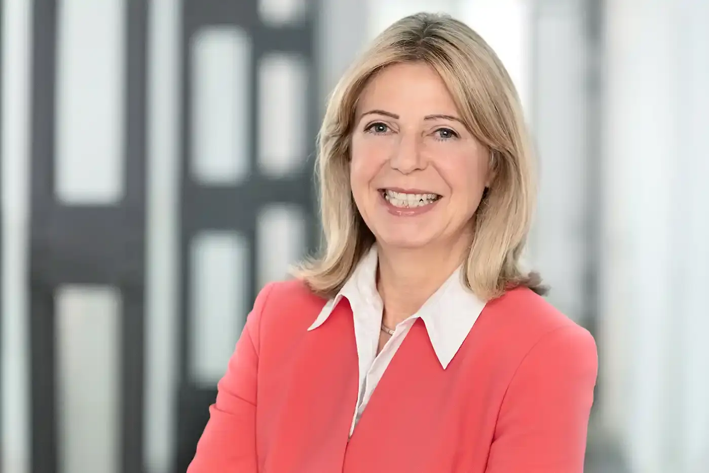 Portraitfoto einer Frau mit blonden langen Haaren, weißer Bluse und einer korallroten Jacke.