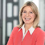 Portraitfoto einer Frau mit blonden langen Haaren, weißer Bluse und einer korallroten Jacke.