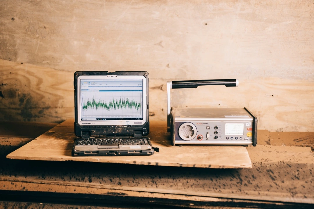 Ein Notebook und ein kastenförmiges Messgerät mit einem Griff stehen nebeneinander auf einem Stapel Baumaterial. Auf dem Notebook sind Messergebnisse in einem Diagramm zu sehen.
