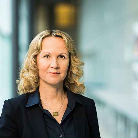 Portraitfoto einer Frau mit blonden gelockten Haaren, die eine dunkle Bluse unter einem Blazer und eine Halskette trägt. Der Hintergrund ist unscharf.