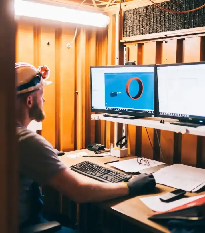 Ein Mann in Arbeitsbekleidung und Helm sitzt in einem gelben Container am Schreibtisch vor zwei Computerbildschirmen. 
