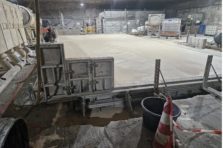 Das Bild zeigt einen Raum im Bergwerk. In der Mitte des Bildes ist eine große Fläche aus frisch gegossenem Beton zu erkennen. Die Fläche ist umgeben von Schalungselementen aus Metall. Um die Fläche herum und im Hintergrund stehen technische Geräte sowie einzelne Lagerbehälter.