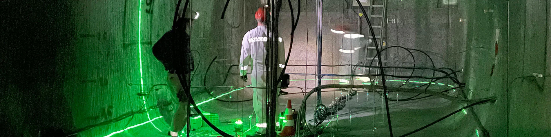 Zwei Personen stehen in einem Bergwerk unter Tage. Um sie herum befinden sich zahlreiche Kabel und Schläuche.