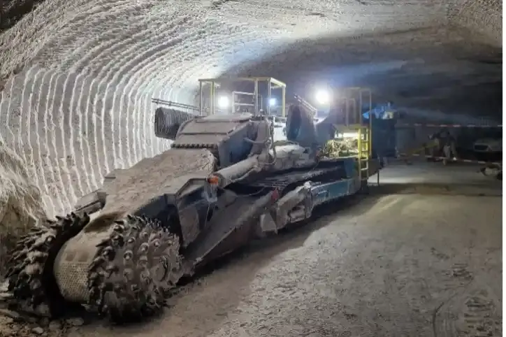 Das Bild zeigt eine große Fräsmaschine in einer Strecke im Bergwerk. Im Hintergrund richten Bergleute einen Arbeitsbereich ein. An der Wand aus Steinsalz sind typische Frässpuren zu erkennen.