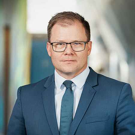 Portrait eines Mannes mit kurzen braunen Haaren und dunkler Brille. Er trägt einen blauen Anzug und Krawatte. Der Hintergrund ist unscharf.