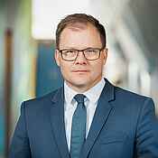 Portrait eines Mannes mit kurzen braunen Haaren und dunkler Brille. Er trägt einen blauen Anzug und Krawatte. Der Hintergrund ist unscharf.