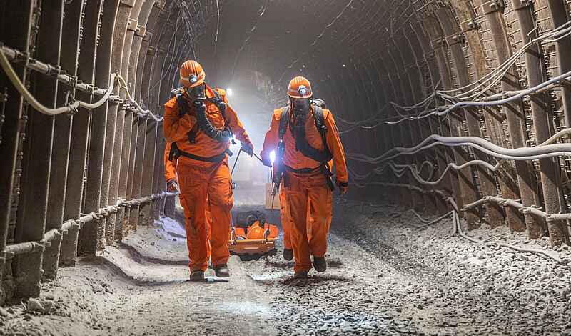 Zwei Personen mit Atemmasken, Helm und Schutzkleidung gehen während einer Übung durch einen Tunnel in einem Bergwerk. Hinter sich ziehen sie eine Art Bahre, auf der eine verletzte Person zu liegen scheint.