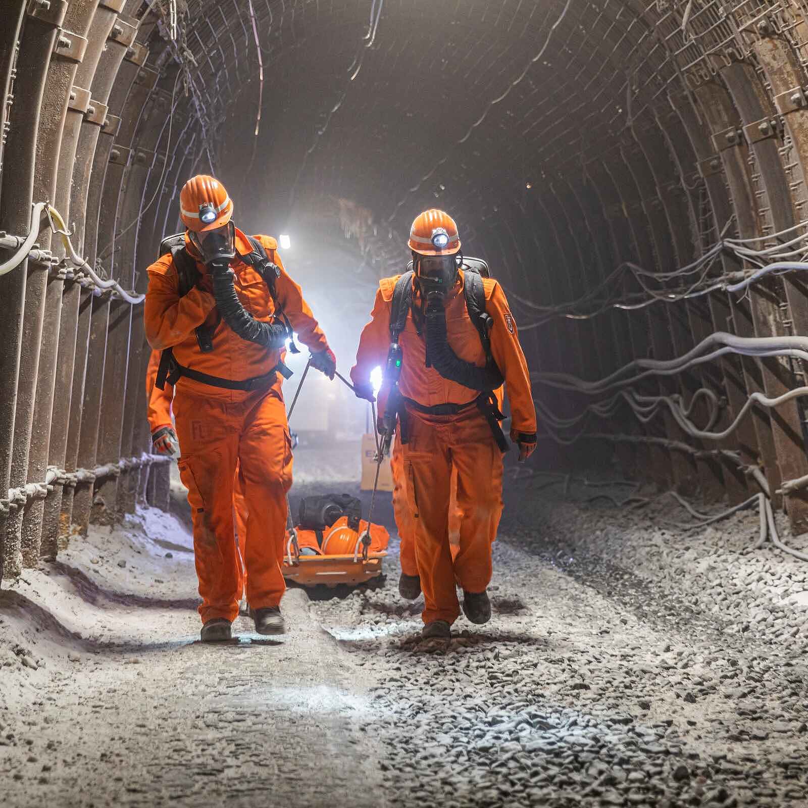 Zwei Personen mit Atemmasken, Helm und Schutzkleidung gehen während einer Übung durch einen Tunnel in einem Bergwerk. Hinter sich ziehen sie eine Art Bahre, auf der eine verletzte Person zu liegen scheint.