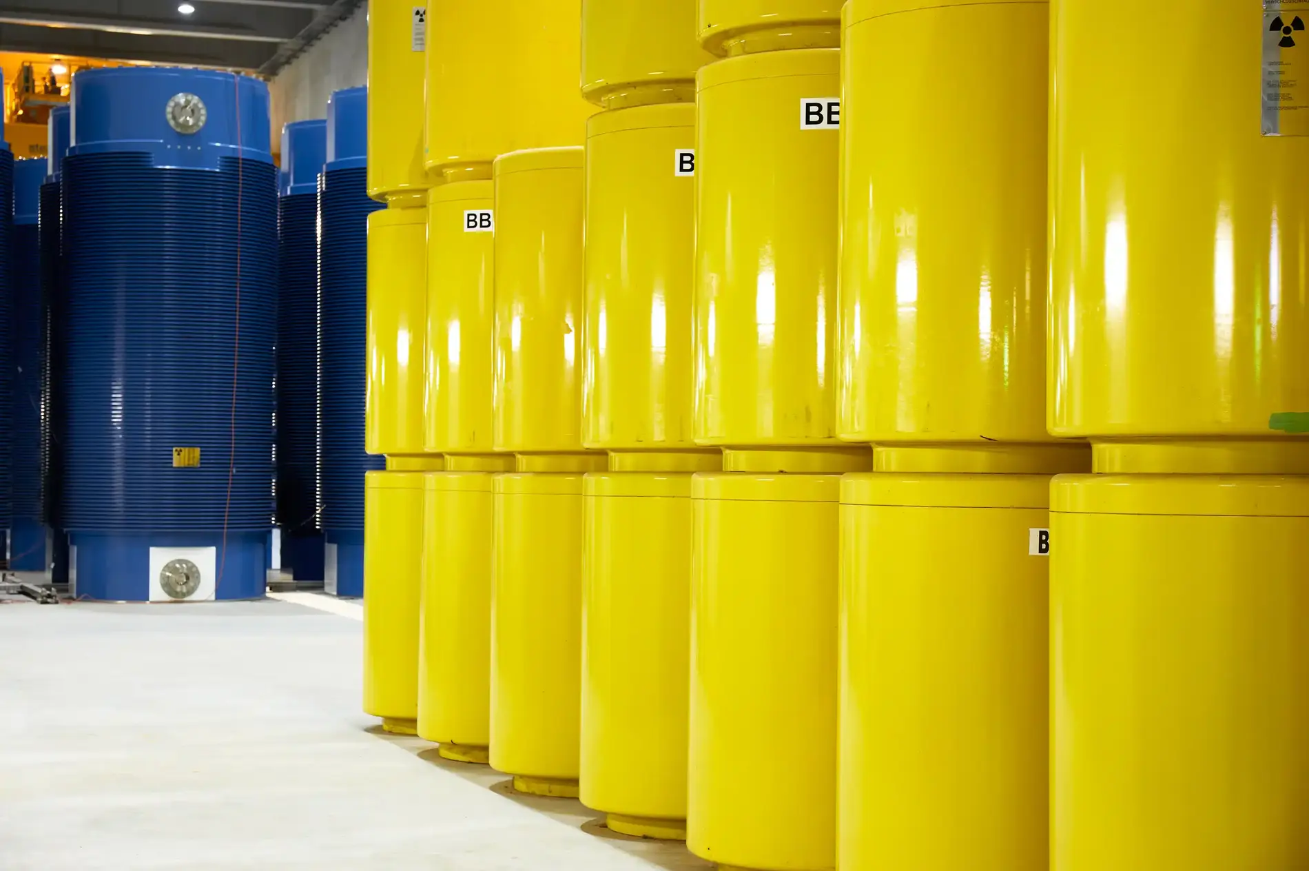 In einer Halle stehen rechts im Vordergrund gestapelte gelbe Fässer. Links im Hintergrund stehen blaue Behälter.
