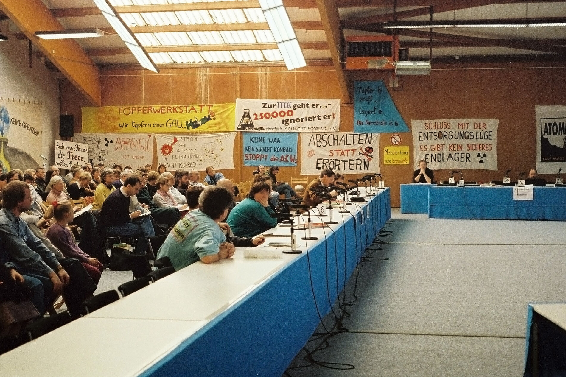 Eine Gruppe von Personen sitzt in einer Halle mit Protest-Transparenten im Hintergrund