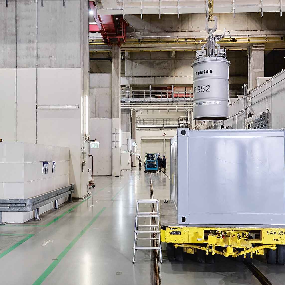Eine Person in Arbeitskleidung steht in großer Halle und bedient mit einer Fernbedienung einen Kran, um ein Fass in einen großen Container zu setzen. 