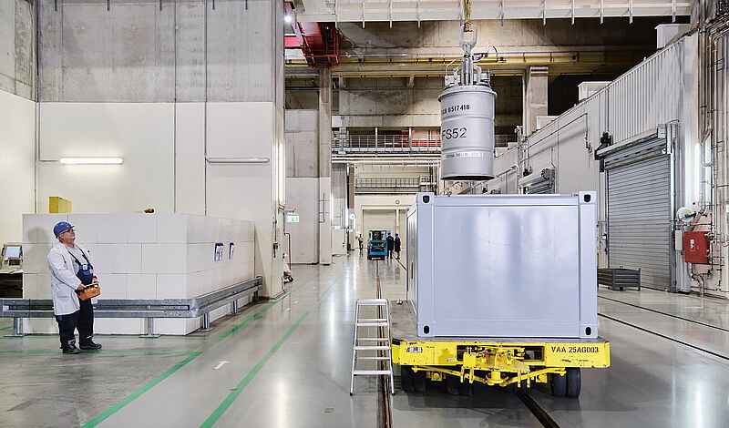 Eine Person in Arbeitskleidung steht in großer Halle und bedient mit einer Fernbedienung einen Kran, um ein Fass in einen großen Container zu setzen. 