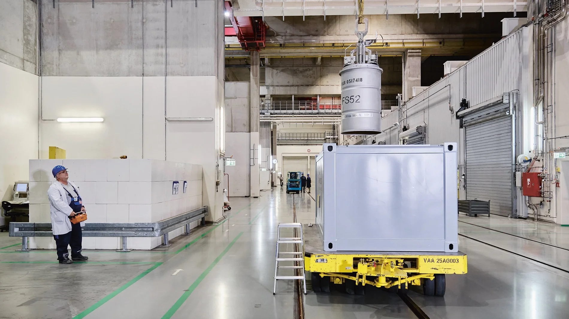 Eine Person in Arbeitskleidung steht in großer Halle und bedient mit einer Fernbedienung einen Kran, um ein Fass in einen großen Container zu setzen. 
