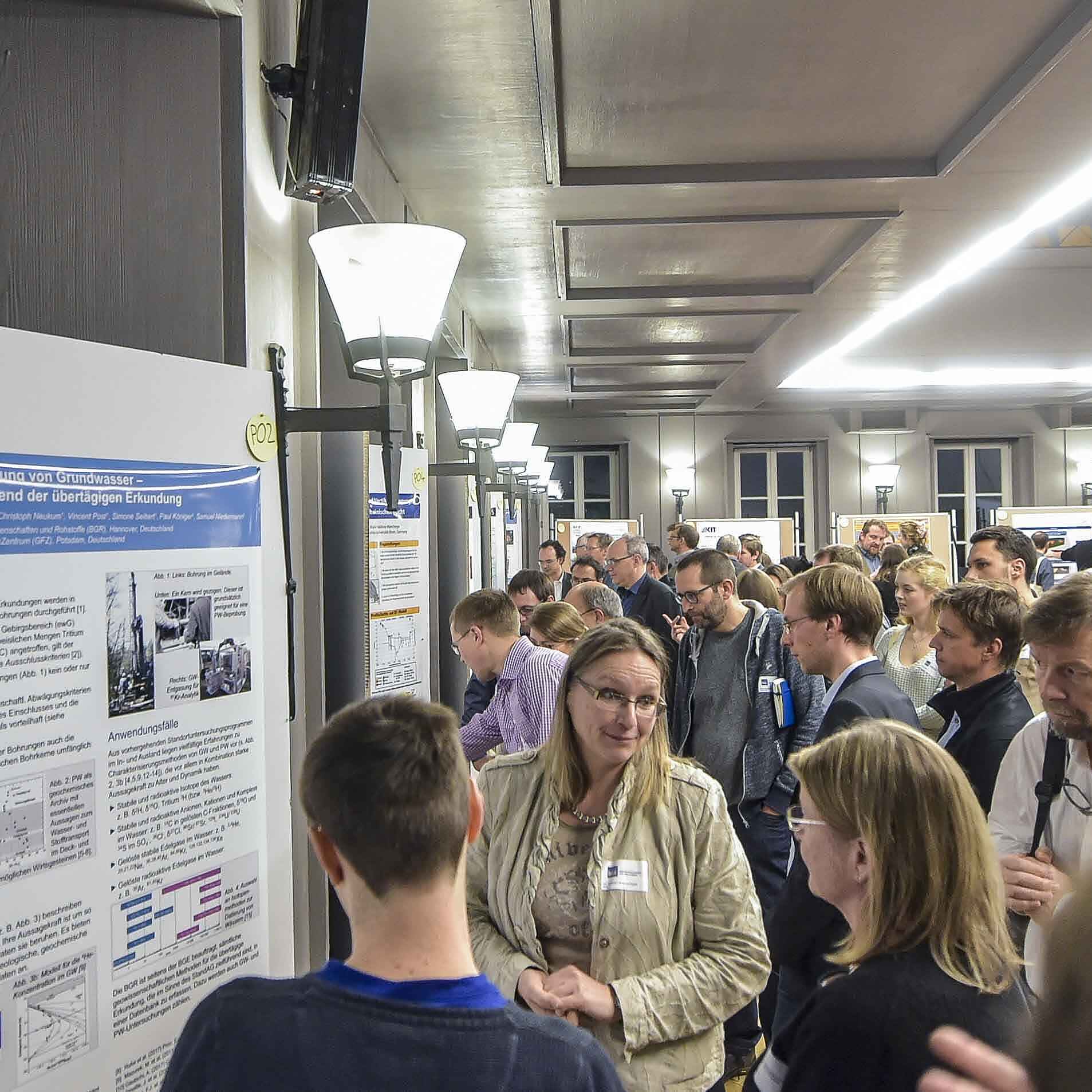 Auf dem Foto ist eine Menschengruppe abgebildet, die sich wissenschaftliche Poster auf einer Pinnwand anschauen und im Gespräch ist. Im Hintergrund sind mehr Menschen und Poster in dem großen Raum zu sehen.