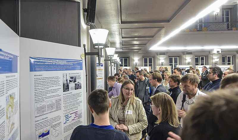 Auf dem Foto ist eine Menschengruppe abgebildet, die sich wissenschaftliche Poster auf einer Pinnwand anschauen und im Gespräch ist. Im Hintergrund sind mehr Menschen und Poster in dem großen Raum zu sehen.