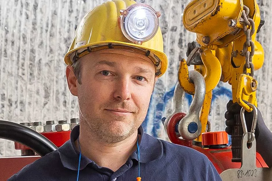 Ein Mann mit gelbem Schutzhelm und blauem Polo-Shirt. Im Hintergrund sind Gestein unter Tage und Arbeitsmaschinen zu sehen.