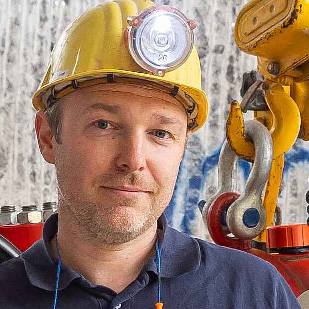 Ein Mann mit gelbem Schutzhelm und blauem Polo-Shirt. Im Hintergrund sind Gestein unter Tage und Arbeitsmaschinen zu sehen.