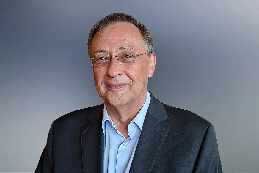 Porträt eines Mannes mit Brille in dunklem Sakko und hellblauem Hemd vor neutral grauem Hintergrund.