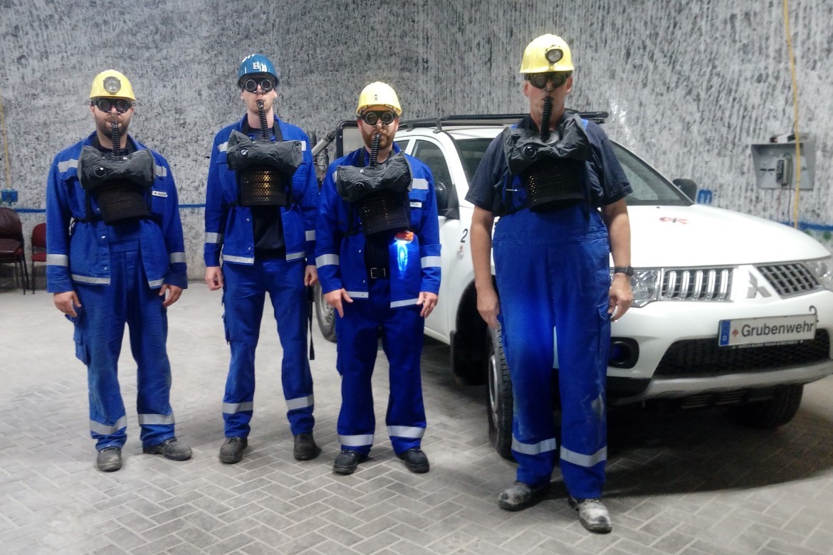 Vier Bergleute stehen mit angelegtem Sauerstoffselbstretter unter Tage vor einem Fahrzeug