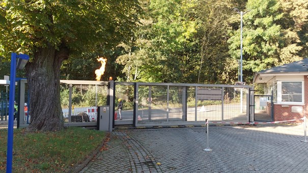 Zu sehen ist das Tor der Schachtanlage, vor dem Tor ein Bauzaun um ein übermannshohes Rohr, an dessen oberem Ende eine etwa mannshohe Flamme brennt.