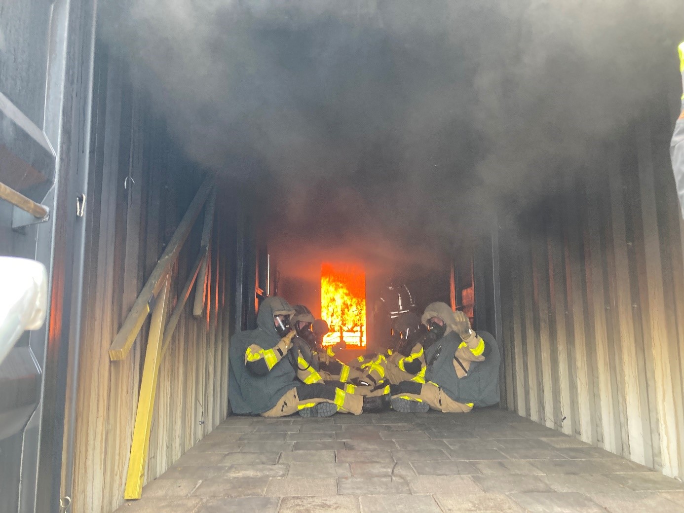 Feuerwehrleute sitzen in voller Schutzkleidung und mit Schutzmasken auf dem Boden eines Containers. Hinter ihnen brennt ein Feuer. Über ihnen steht starker Rauch. 