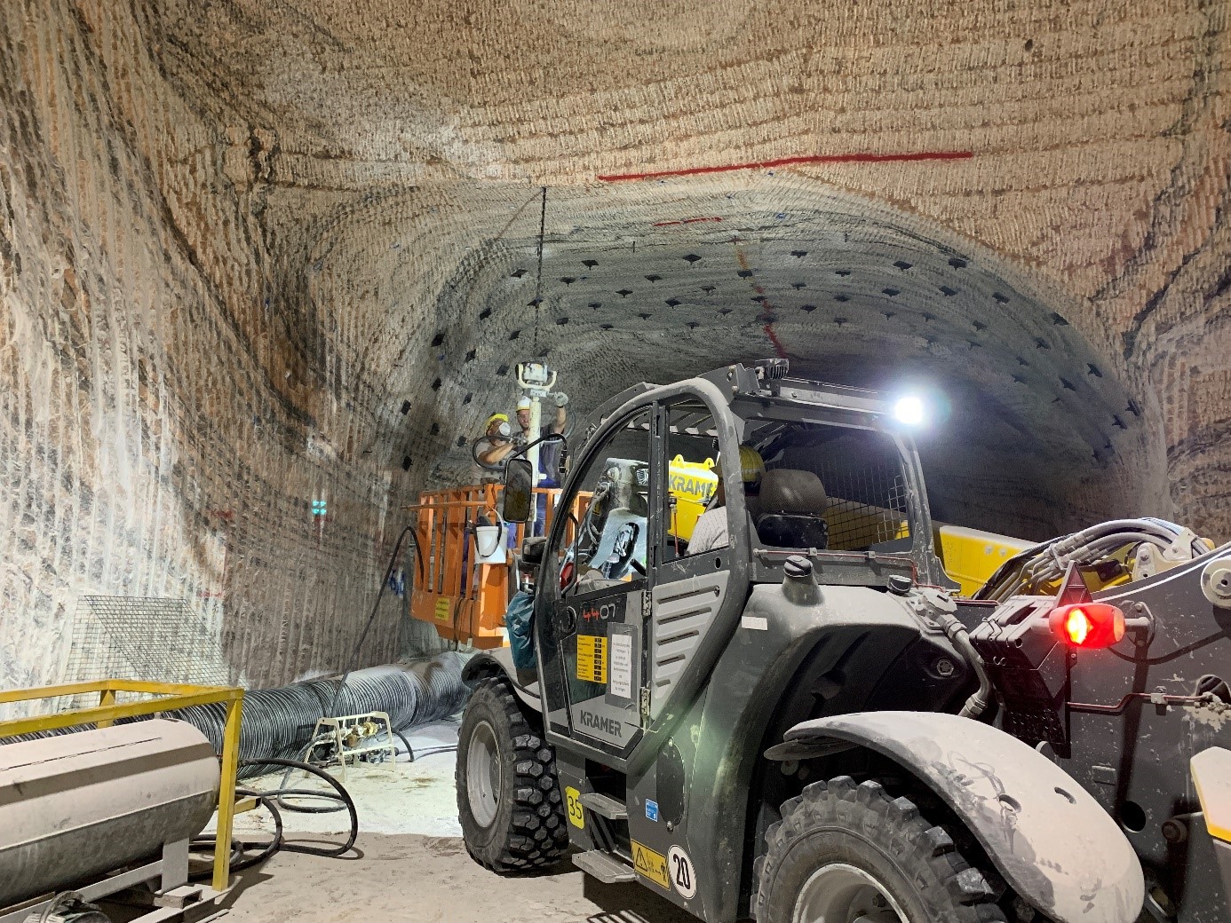 Zwei Bergleute stehen auf einer Hebebühne eines Fahrzeugs. Sie bedienen einen großen Bohrer, um ein Bohrloch in die Decke zu stoßen. Hinter den Mitarbeitern stecken Anker in der Decke, die durch ein Netz miteinander verbunden sind.