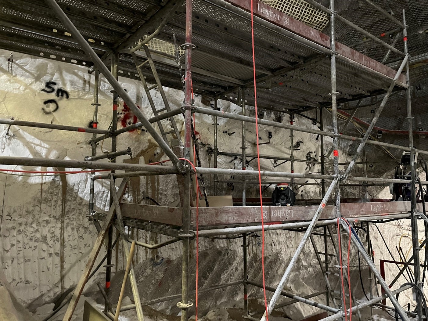 Ein Baugerüst steht direkt vor einer Salzwand unter Tage. In der Wand befinden sich Löcher für Bohrungen. In einigen Bohrlöchern befinden sich Metallstäbe, sogenannte Gebirgsanker. Neben den Löchern sind Markierungen gesprüht, die auf die Länge dieser Anker hinweist. Die Markierung ist 5 Meter.