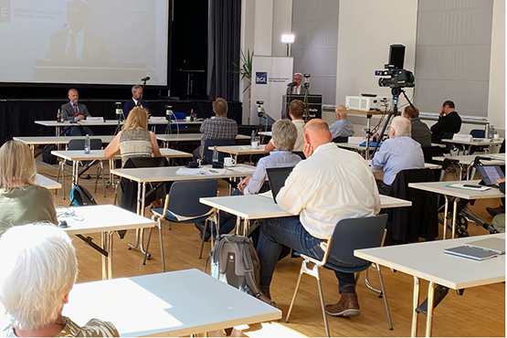 Das Foto zeigt denn Blick in den Veranstaltungsraum. Personen sitzen an Tischen und folgen der Veranstaltung. BGE-Mitarbeiter Lutz Teichmann hält einen Vortrag am Rednerpult.