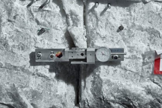 Das Foto zeigt ein Messgerät mit einer runden Anzeige, das mit Schrauben im Gestein verankert ist.