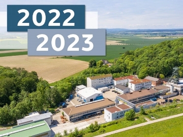 Luftaufnahme von mehreren weißen Gebäuden; grüne Laubbäume und Felder schließen sich daran an. Im Bildrand links oben stehen die Jahreszahlen 2022 und 2023 in großer Schrift.