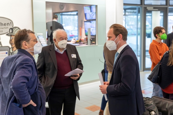 Drei Männer mit FFP2-Masken unterhalten sich im Foyer eines Bürogebäudes.