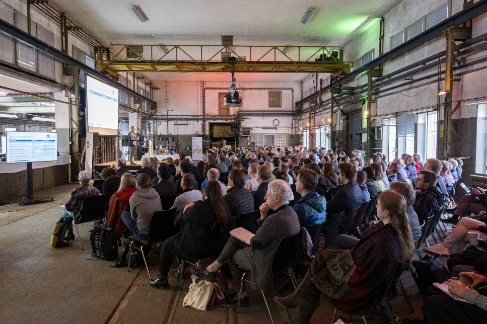 Auf dem Foto sind zahlreiche sitzende Menschen verschiedenen Alters zu sehen, die in einer Halle zur Bühne blicken. 