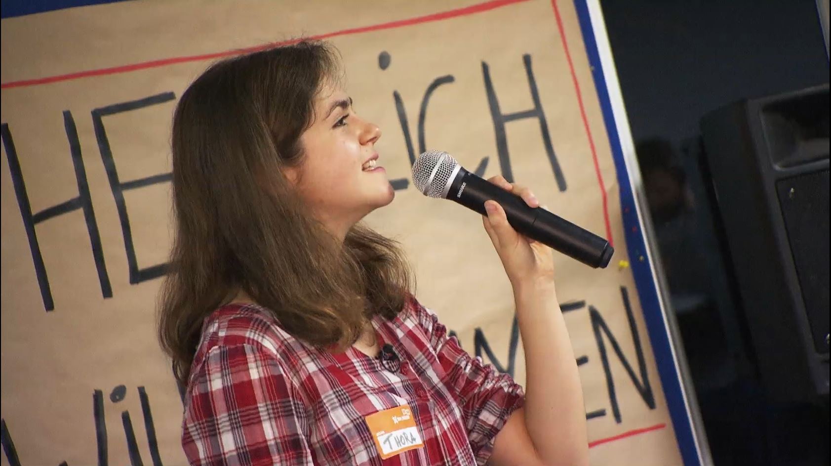 Eine junge Frau in einer karierten Bluse und mit braunen langen Haaren spricht in ein Mikrofon.