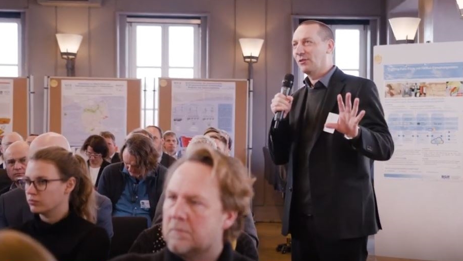 Ein Mann im dunklen Anzug spricht gestikulierend in ein Mikrofon, neben ihm sitzen Menschen im Publikum. Im Hintergrund sind Ausstellungstafeln und Pinnwände mit Plaketen zu sehen.