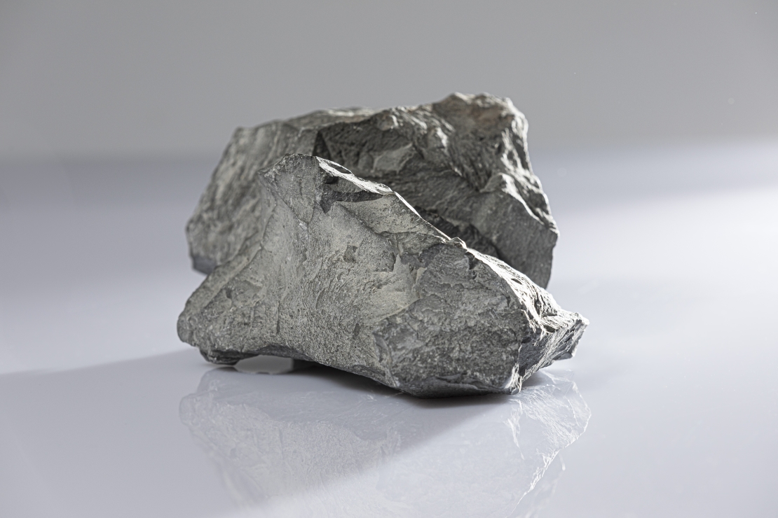 Foto eines dunklen Brockens Tongestein. Der Stein befindet sich auf einer hellen, reflektierenden Oberfläche und wirft einen Schatten nach links aus dem Bild heraus.