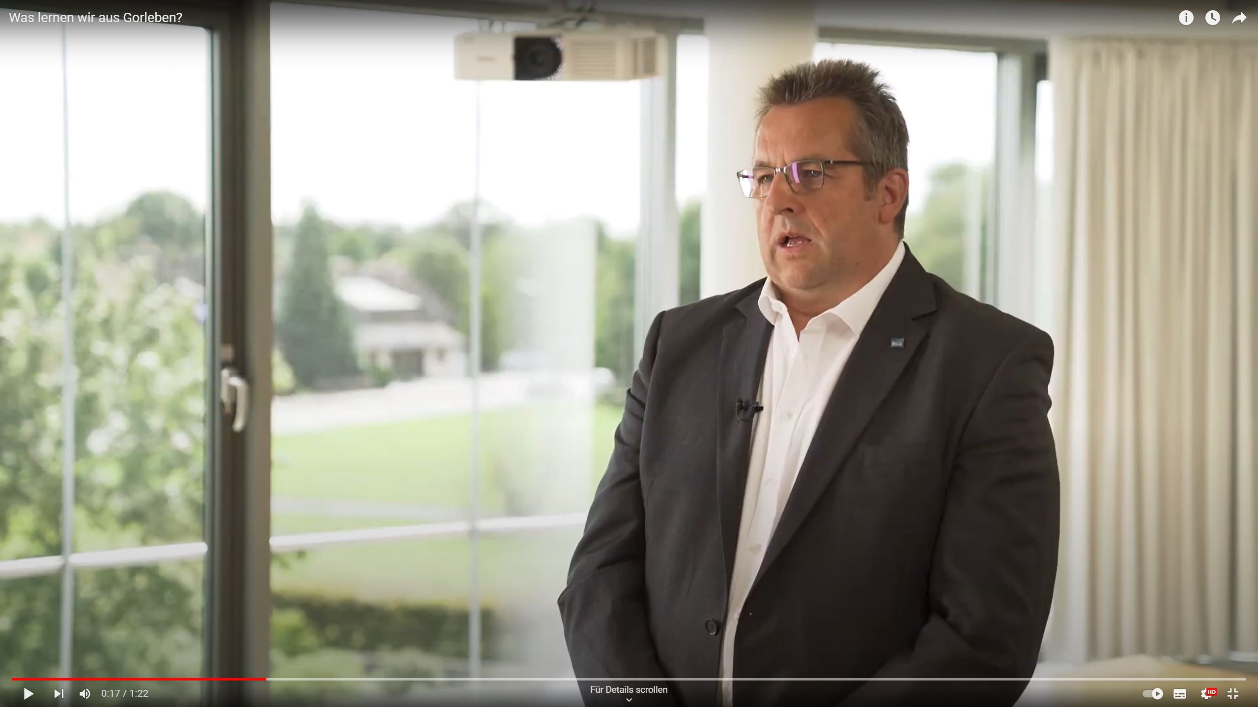 Screenshot aus dem Video, das einen Mann mit Brille, Sakko, weißem Hemd und kurzen graubraunen Haaren vor einer Fensterfront zeigt.