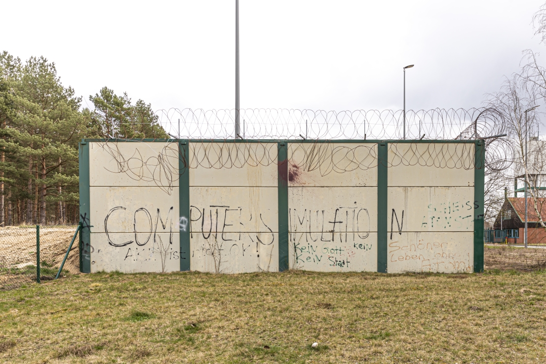 Foto eines isoliert stehenden Stücks einer grau-beigen Mauer mit Graffitis und Stacheldraht.