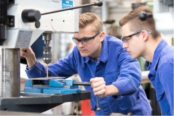 Zwei Auszubildende zum Industriemechaniker bohren ein Loch in eine Metallplatte.