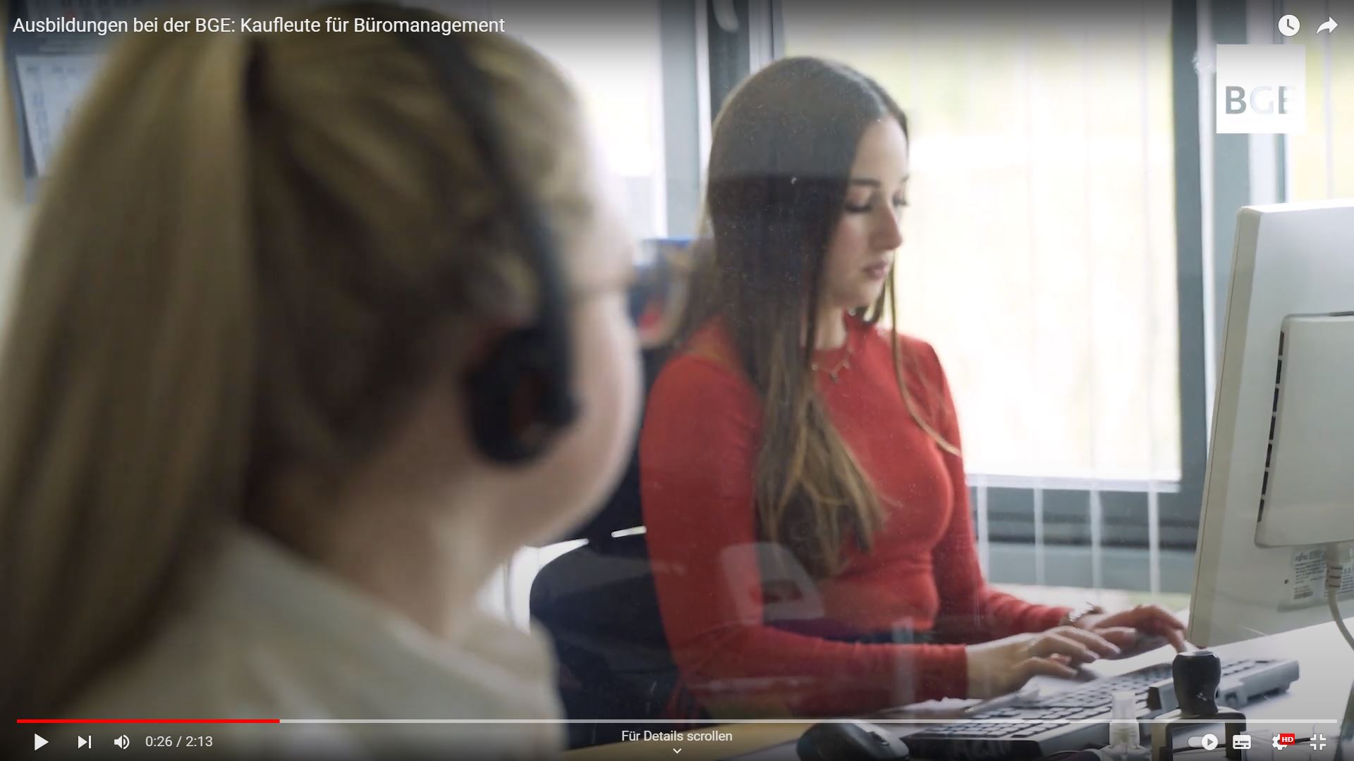 Foto von zwei Frauen. Die Frau im Vordergrund trägt eine Brille und ein Headset. Die Frau im Hintergrund trägt einen roten Pullover und arbeitet an einem Computer