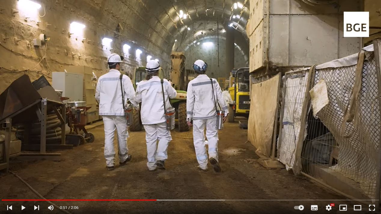 Foto der Rückenansicht von drei Personen in weißer Bergmannskleidung, die eine Strecke unter Tage entlanggehen.