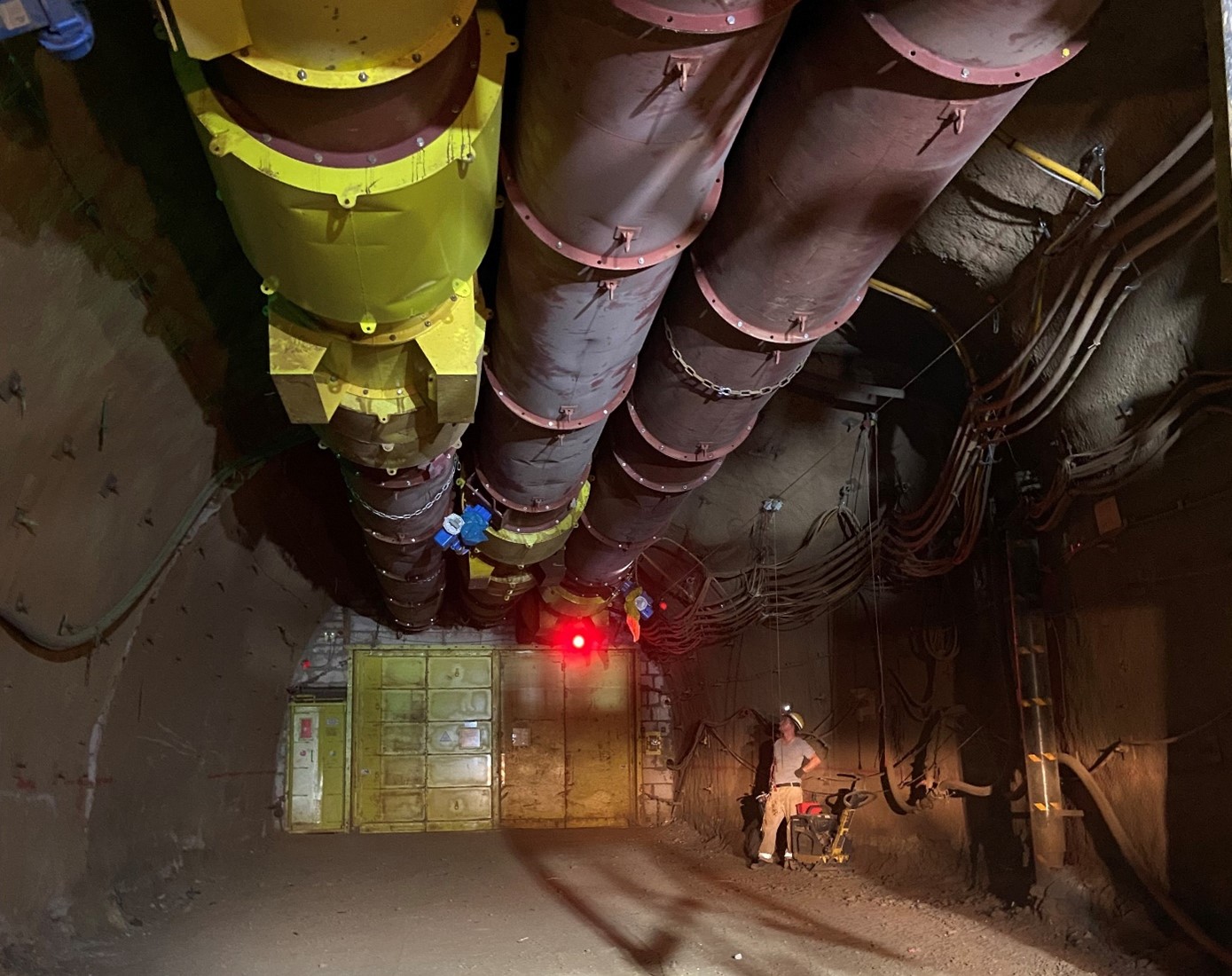 In einen Tunnel hängen drei Große Röhren an der Decke. Am ende des Tunnels ist eine Metallwand. eine Arbeiter steht am Boden und schaut zur Tunneldecke.