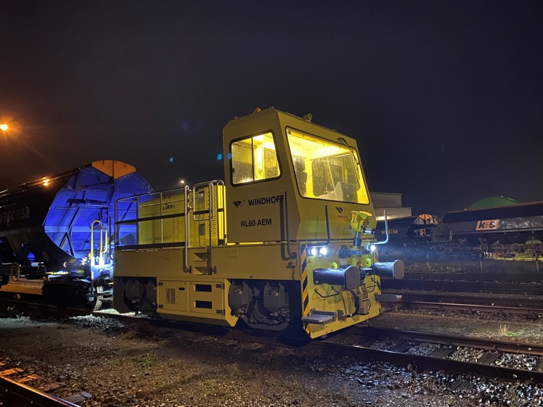Eine gelbe Lokomotive steht in abendlicher Atmosphäre auf Bahnschienen. Das Führerhaus ist von innen hell erleuchtet. Die Lokomotive hat einen blauen Waggon angekoppelt.
