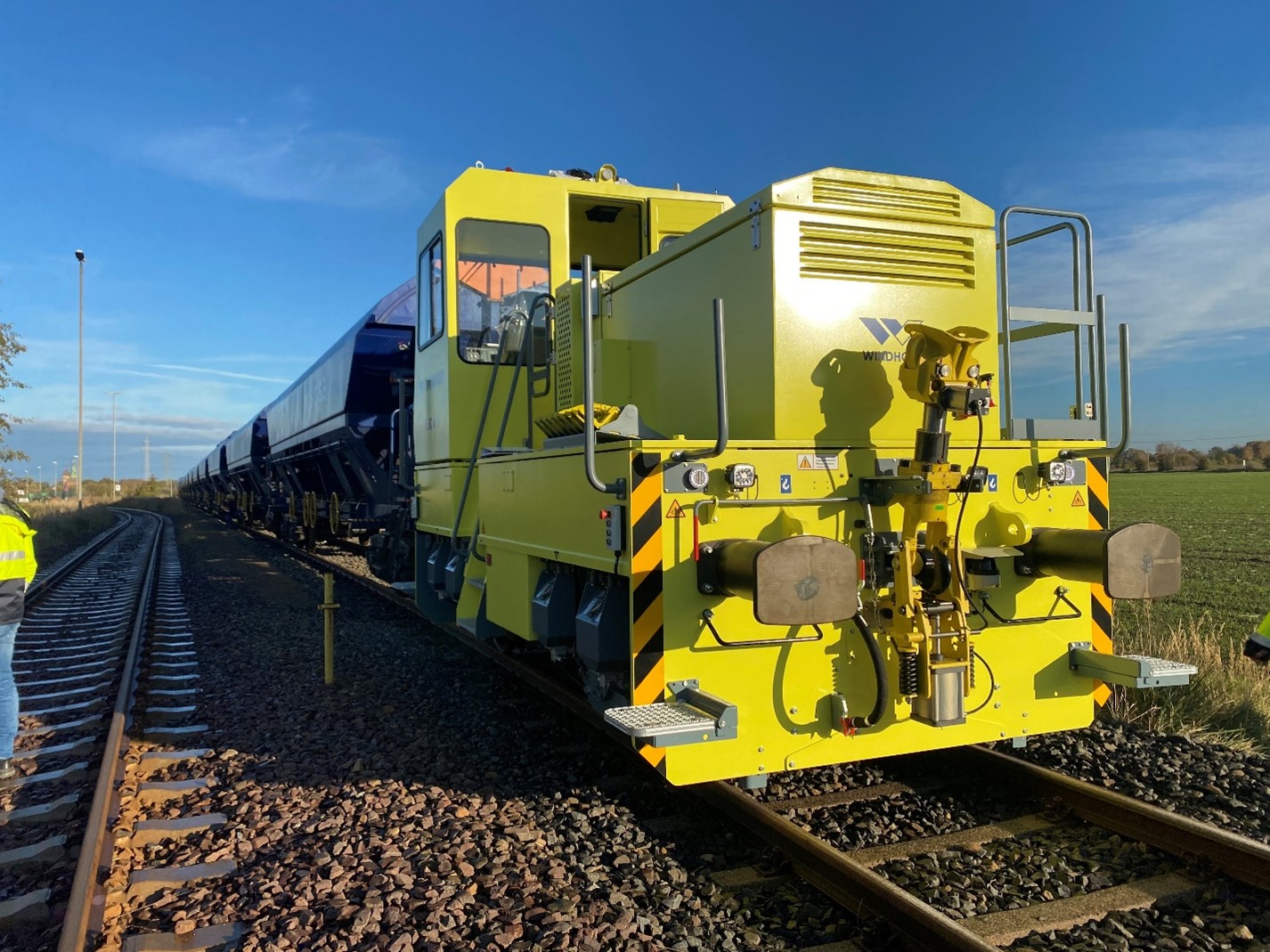 Das Foto zeigt eine leicht schräge Frontalaufnahme einer gelben Lokomotive. Sie steht auf Bahnschienen an einem Feld an einem sonnigen Tag mit blauem Himmel. Die Lokomotive hat mehrere blaue Waggons angekoppelt.