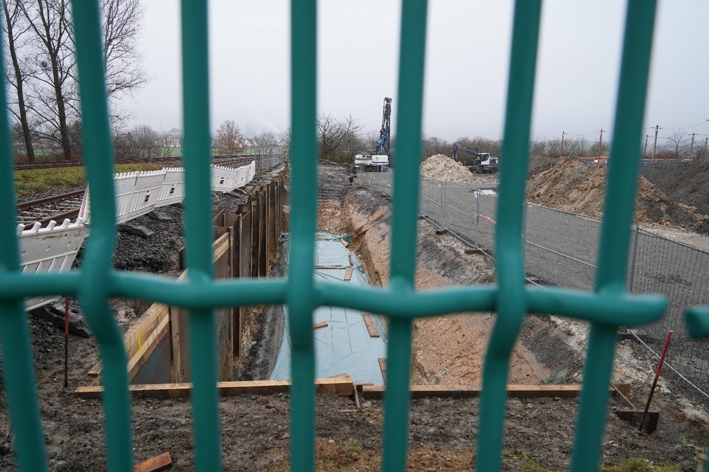 Hinter einem grünen Zaungeflecht erstreckt sich eine längliche Baugrube. Ihr Boden ist mit einer grauen Plane ausgelegt. An der linken Seite wurden Spundwände in den Boden gerammt. Die Baugrube ist von Bauzäunen eingerahmt und im hinteren Bildteil arbeiten zwei Bagger zwischen Sand- und Erdhaufen.