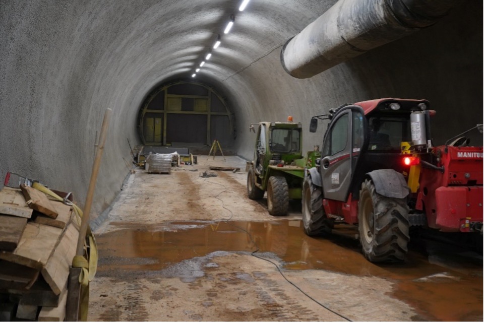 Foto einer Strecke unter Tage, die an einen Tunnel erinnert. Am rechten Bildrand stehen ein grünes und ein rotes Baustellenfahrzeug. Auf dem Boden befindet sich eine große, flache Pfütze. Am linken Bildrand sind Holzbretter aufgestapelt. Mittig an der Decke installierte Leuchtstoffröhren illuminieren die Strecke.