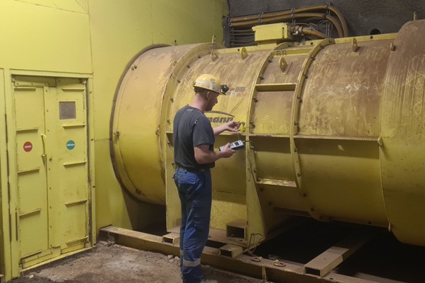 Ein Mann in Arbeitskleidung steht mit einem Messgerät in der rechten Hand vor einer zylindrischen gelben Maschine. Er trägt einen gelben Schutzhelm und hält die Messfühler an die Oberfläche der Maschine.