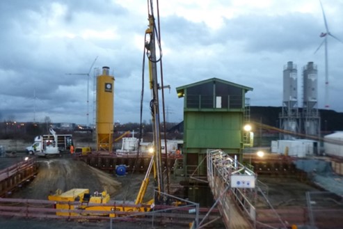 Foto einer Baustelle mit einem gelben Baustellenfahrzeug, Siloanlagen, Lattenzäunen und Bauzäunen. Ein provisorischer Weg führt zu einem grünen Gebäude.