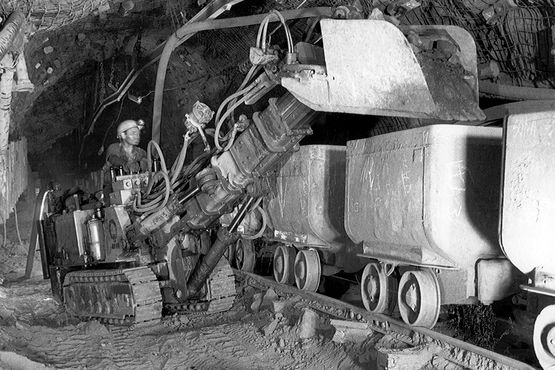 The historic photograph shows a miner loading a mine wagon with ore rock using a shovel loader.