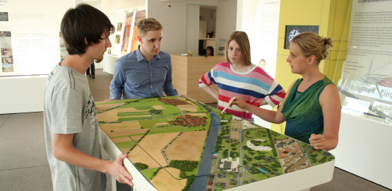 Four people stand around a model of the Konrad mine. Link to page "Info centre and tours"